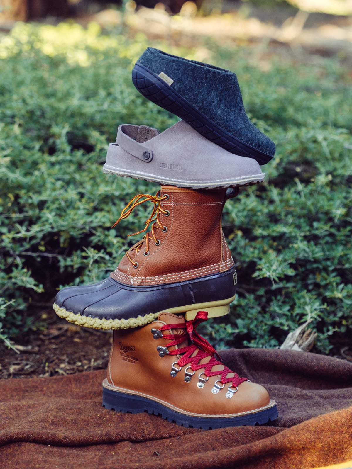 A dark gray wool slipper, a light gray slip-on clog, a brown leather duck boot, and a brown hiking boot with red laces stacked on a brown blanket in front of green foliage.