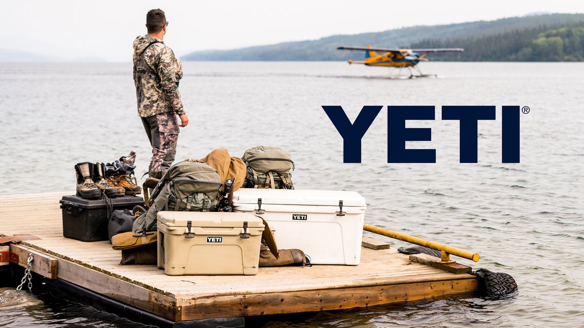 A person in camouflage gear stands on a wooden dock next to YETI coolers and outdoor equipment, with a seaplane visible on a lake surrounded by forested shoreline.