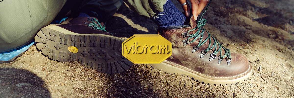 A large Vibram logo embossed on an image of a person wearing brown leather Danner hiking boots who sits on a dirt trail, the treads of their shoes revealing the yellow octagonal Vibram logo.