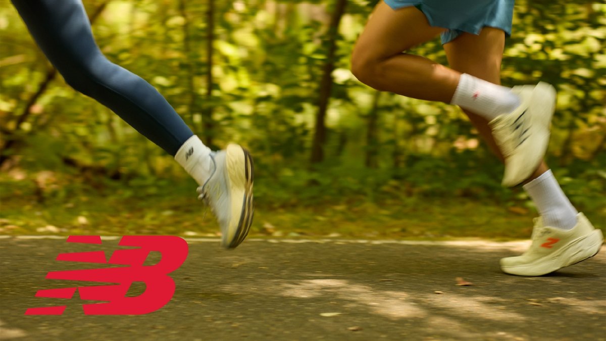 A close-up, motion-blurred shot of two runners' legs and New Balance shoes on a paved path in a forest.