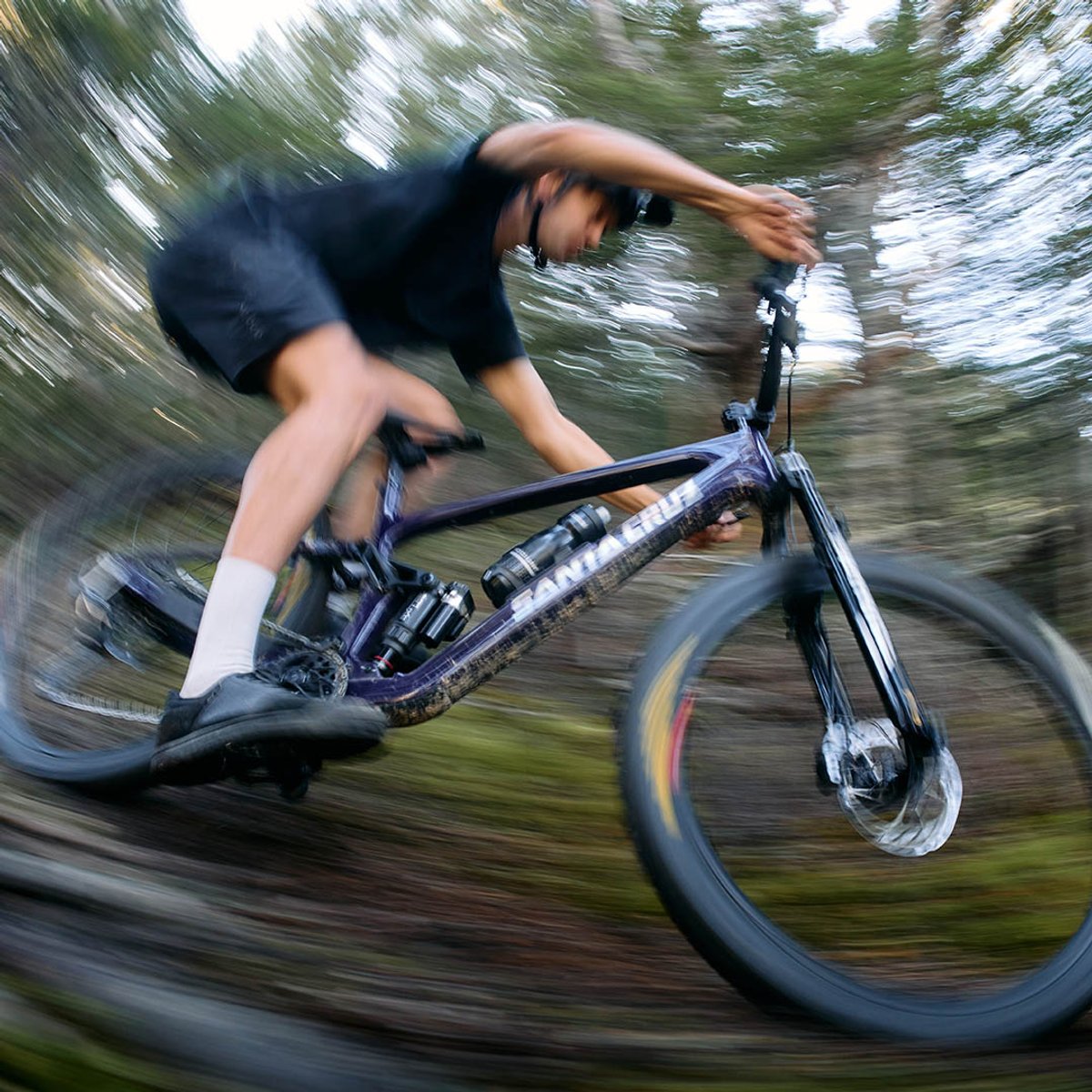 A rider leans hard into a berm on a purple Santa Cruz mountain bike, captured with a radial motion blur that emphasizes speed through a forested trail