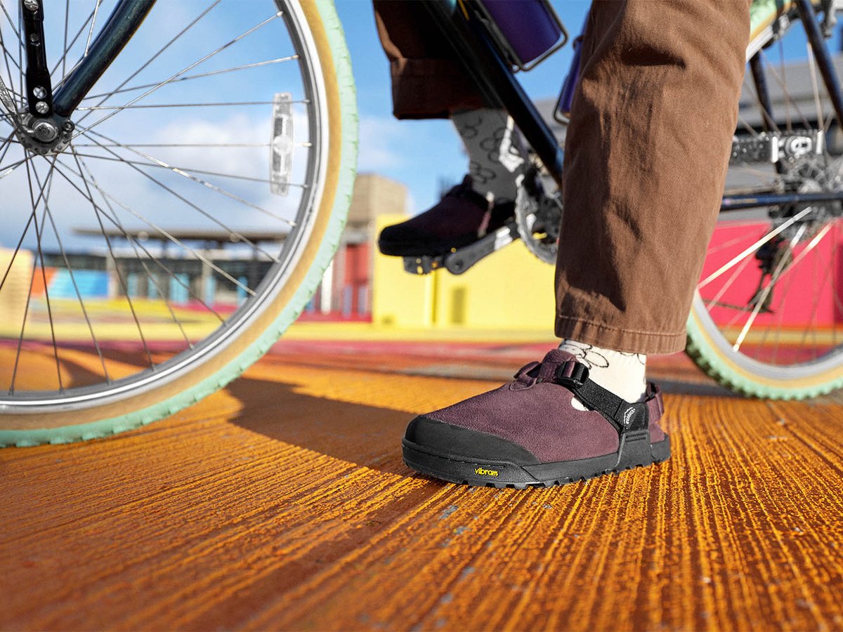 Close-up of a foot in a plum suede Bedrock Mountain Clog with white socks beside a bicycle wheel on a textured orange surface