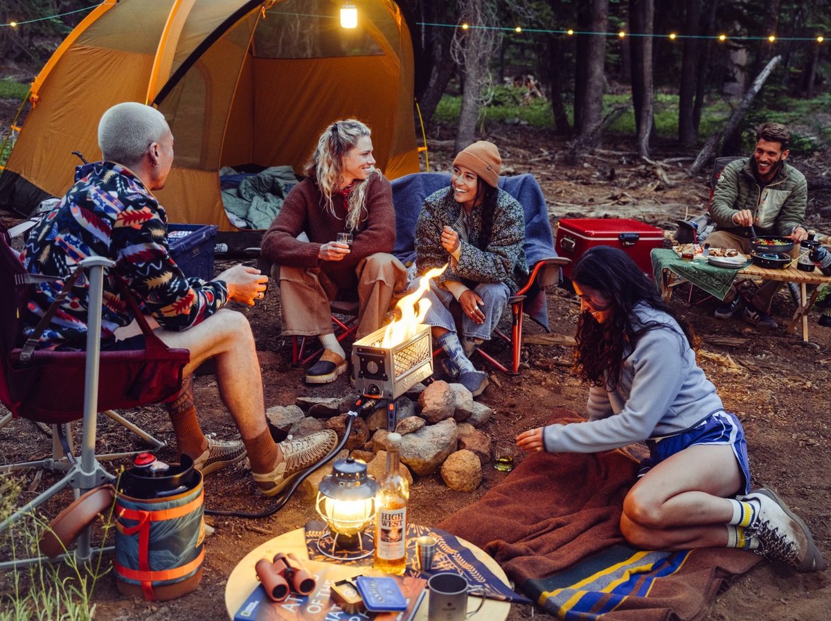 Five friends gathered around a small camp stove fire in a pine forest, laughing and sharing whiskey beside a glowing orange tent.