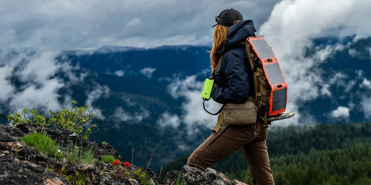 A hiker climbs a rocky ridge with a Dark Energy Spectre folding solar panel strapped to her pack, charging a hi-vis green Poseidon power bank, with clouds and forested mountains behind her.