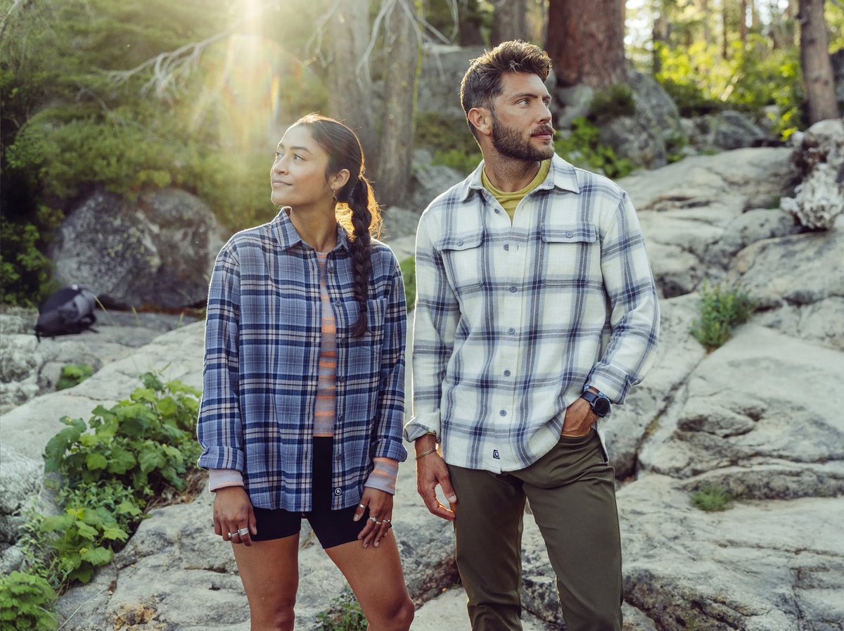 Two people in plaid flannel shirts stand on a granite outcrop surrounded by forest, sunlight filtering through the trees behind them.