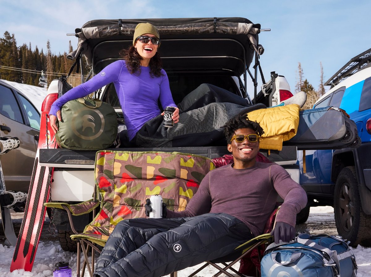 Two friends tailgating from a truck bed at a snowy ski resort, laughing and relaxing in base layers with gear bags and canned drinks nearby.