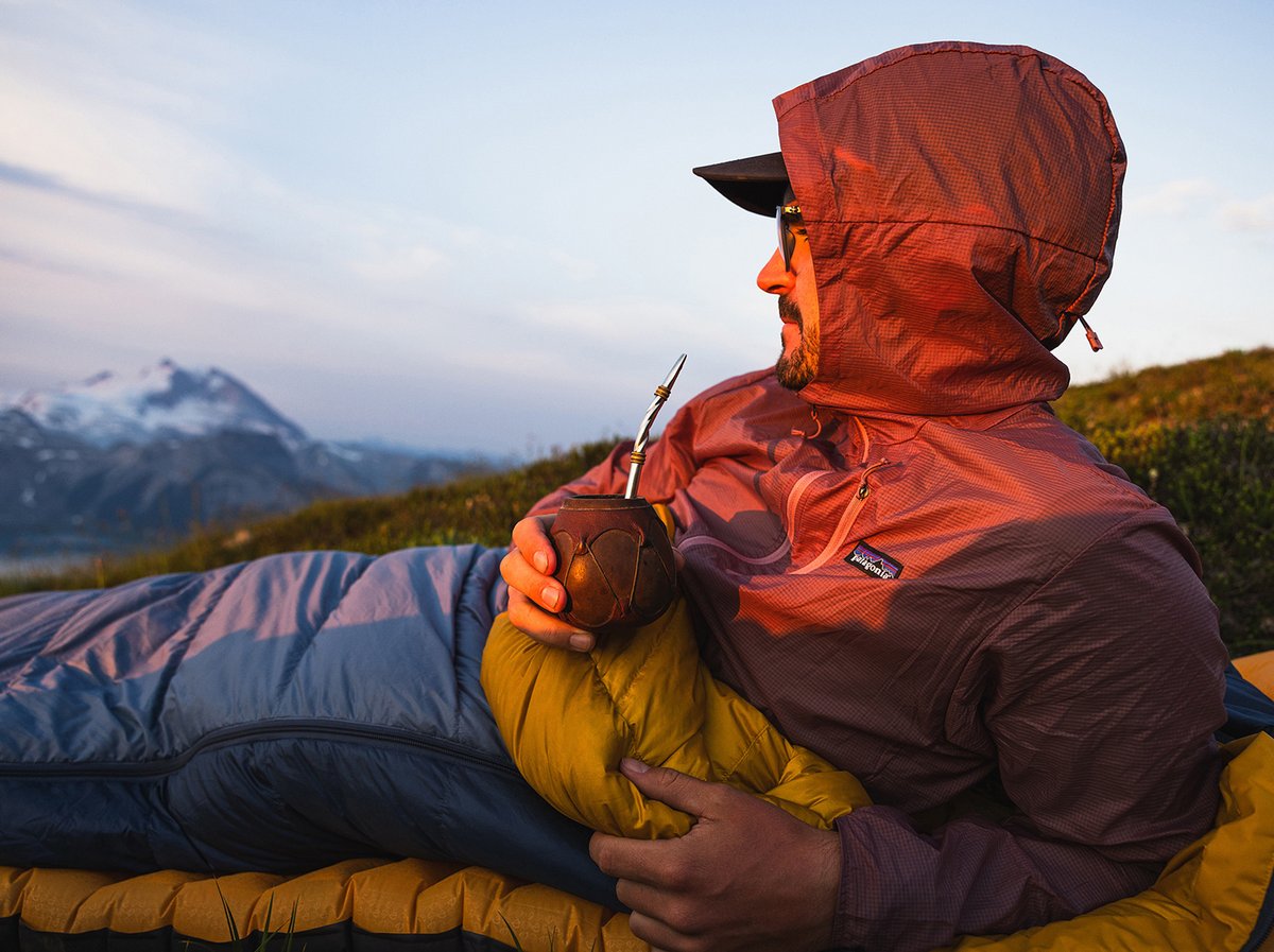 A man sips a drink while laying down in his sleeping bag.