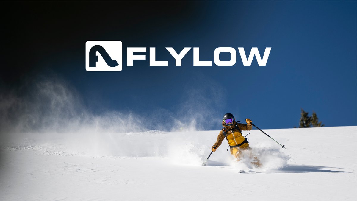 A skier in a yellow and brown technical suit carves through deep powder on a snowy mountain slope under the Flylow logo.
