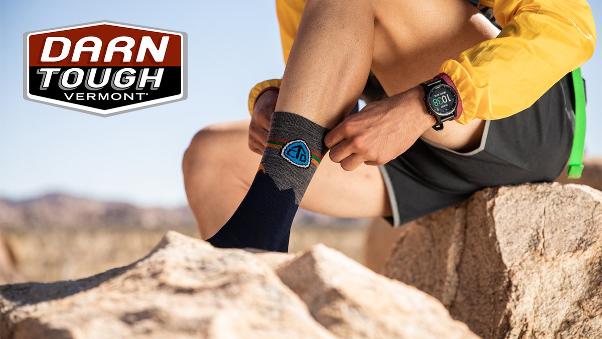 A hiker sitting on a rock in a desert landscape adjusts their Darn Tough Vermont socks, which feature a mountain design.