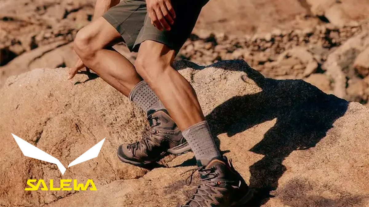 A hiker wearing Salewa brown hiking boots and gray socks stands on rocky desert terrain, showcasing durable outdoor footwear for mountain and trail adventures.