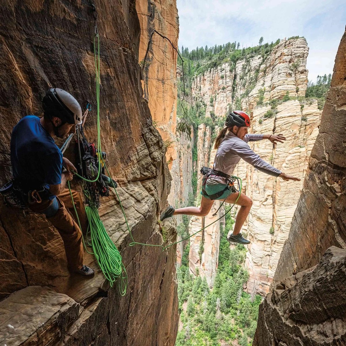 A woman and man are rock climbing on the side of a sheer rock face. The woman is mid-leap to the other side of the rock face.