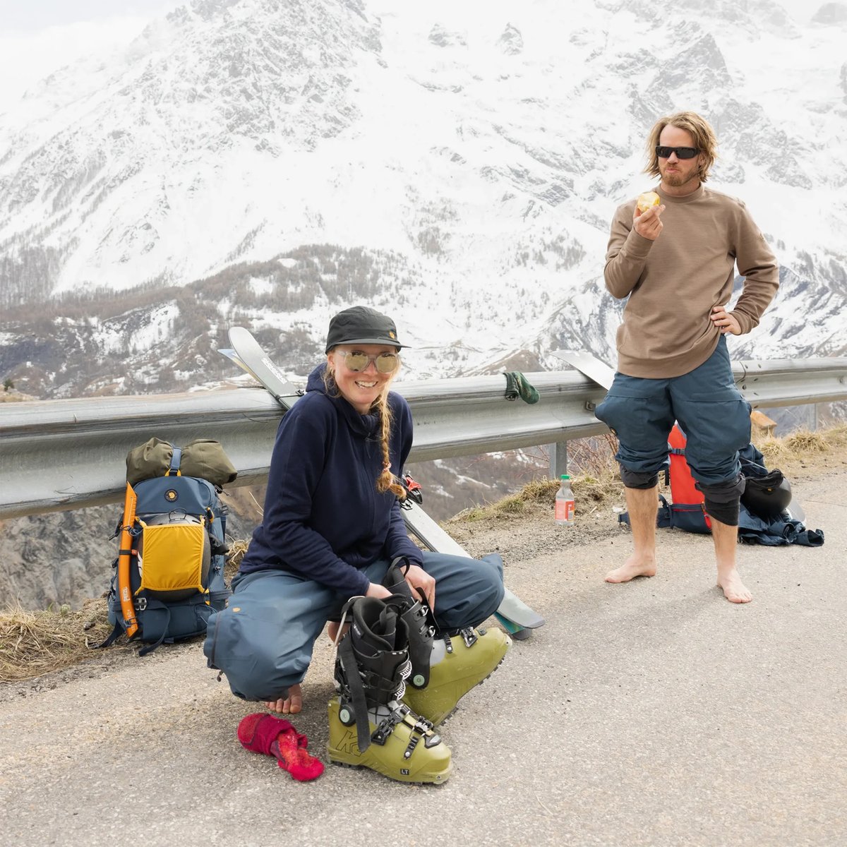 Two hikers taking a break at a mountain overlook, with one wearing ski boots and holding ski equipment while eating a snack, showcasing alpine outdoor adventure gear.