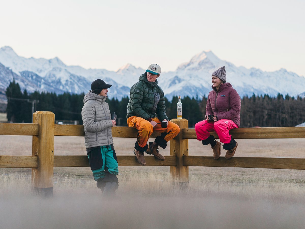 Three people in insulated jackets and ski pants gathered around a wooden fence at dusk with snow-capped peaks in the background.
