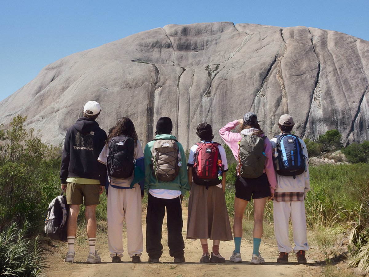 Six people wearing a variety of The North Face backpacks facing a massive granite dome under a clear blue sky.