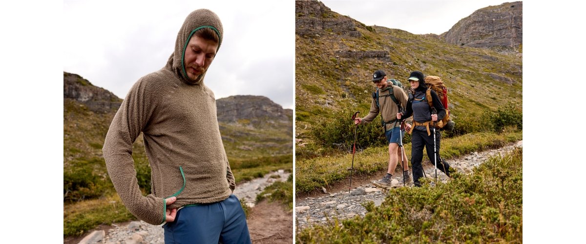 A person stands outdoors on a rocky mountain trail wearing a textured brown hoodie and blue shorts. In the background, two other people equipped with backpacks and trekking poles hike together through grassy, mountainous terrain. The sky is overcast and the landscape features rolling hills and rocky slopes.