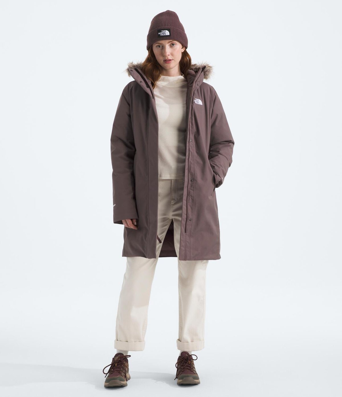 A woman wearing a hat and Arctic Down Parka stands against a neutral background