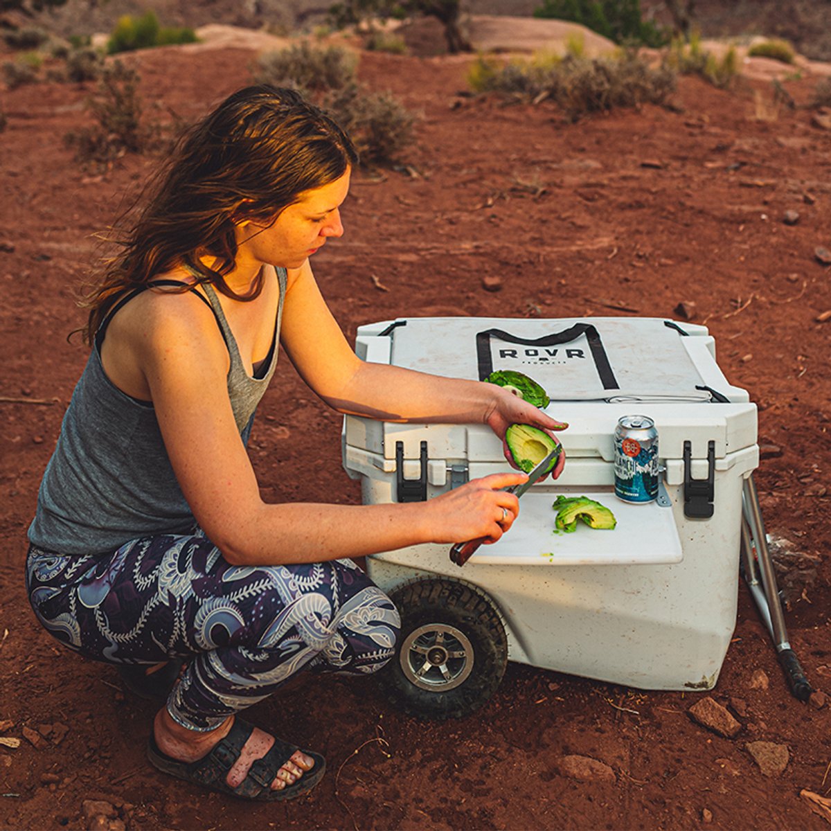 RovR RollR wheeled cooler used as a prep surface while camping, with food and a drink placed on top in a desert setting.
