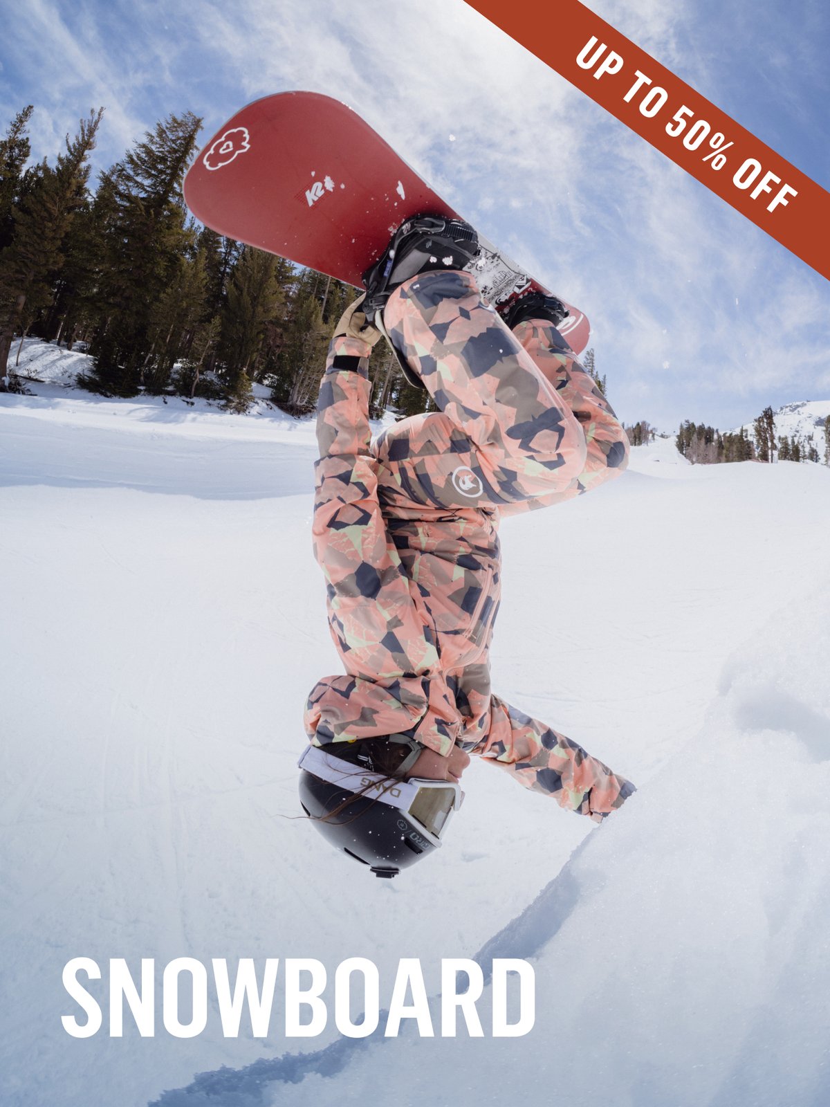 Snowboarder mid-invert on a sunny groomer, holding a red K2 board overhead, wearing a coral camo one-piece suit. Snowboard Up to 50% off.