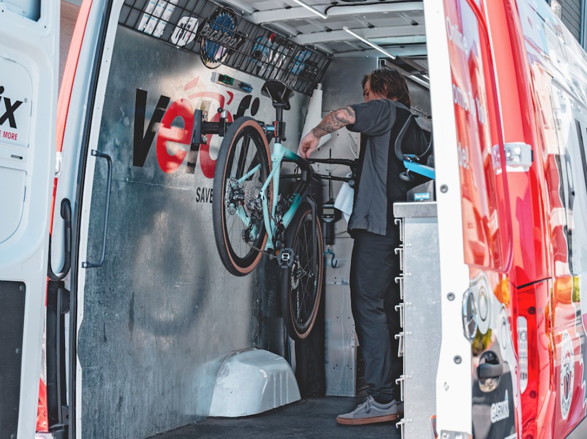 A tattooed mechanic works on a mint-green gravel bike mounted inside a branded mobile bike service van.