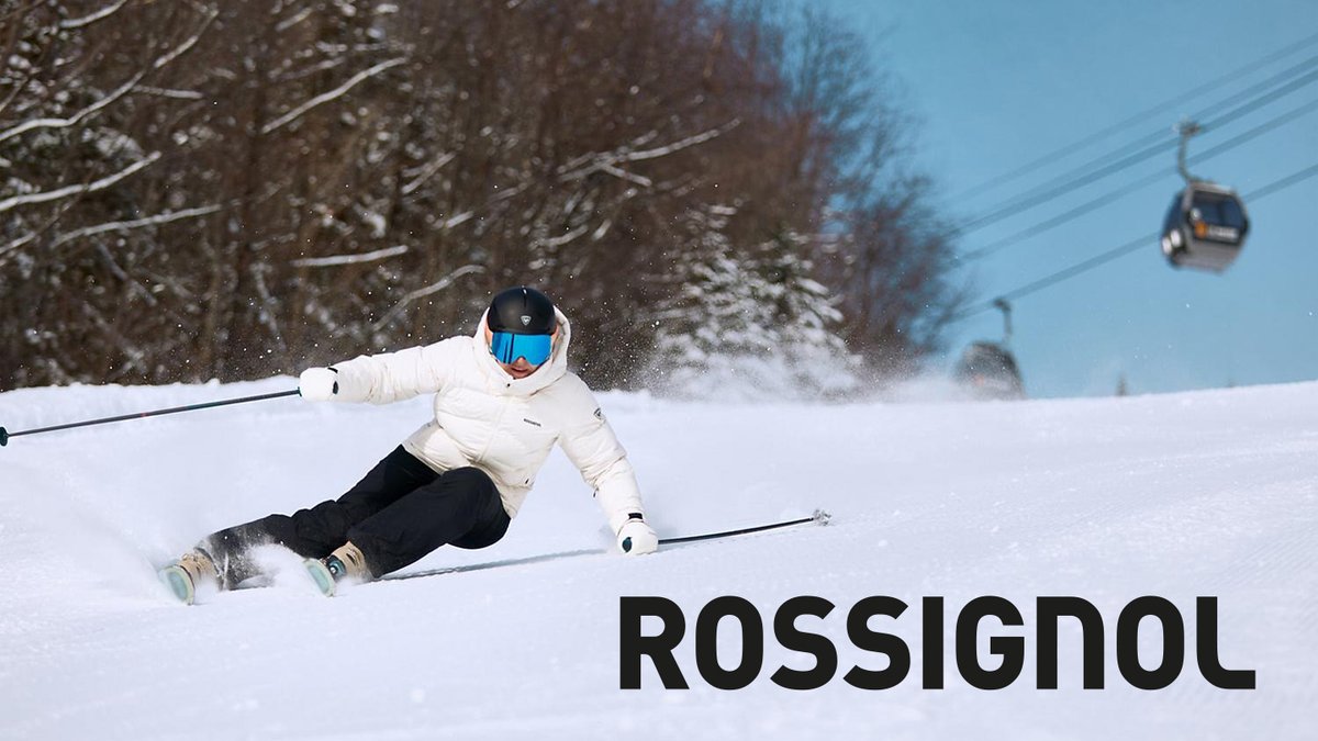 A skier in a white jacket and black pants carves through snow on a groomed slope under a ski lift.