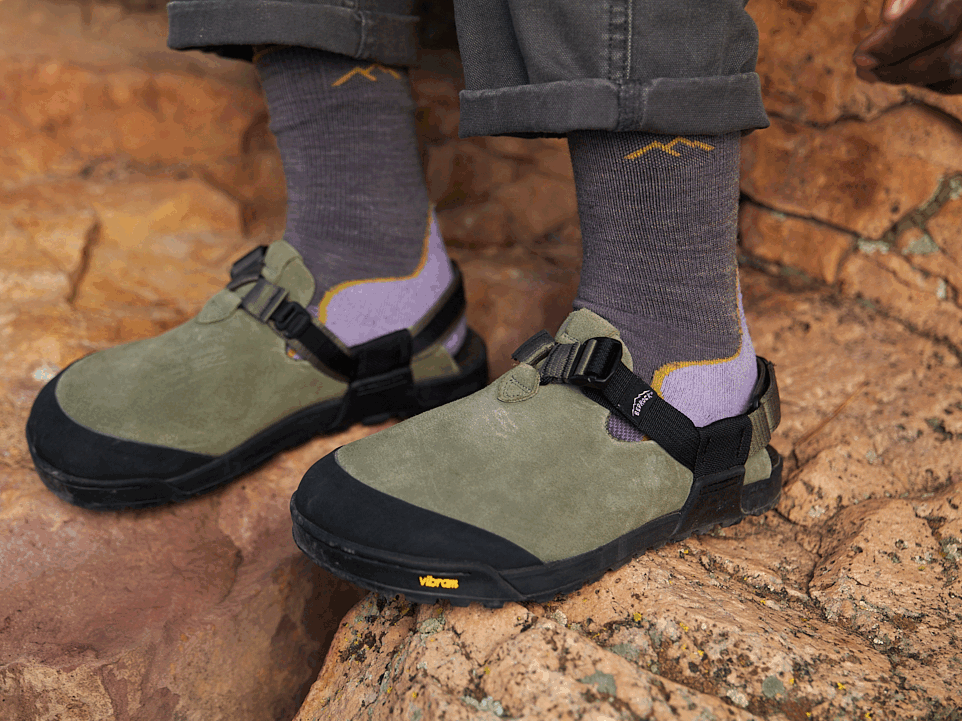 Close-up of hands adjusting heel strap on Bedrock Mountain Clog in olive suede with layered socks on red rock terrain