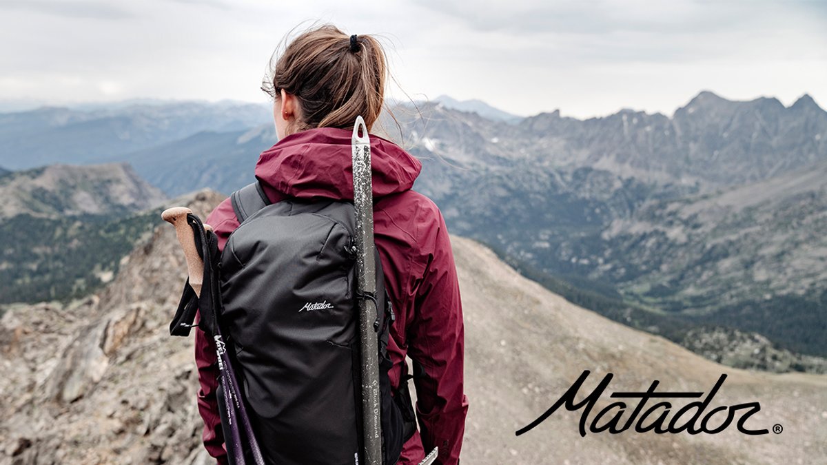 A hiker wearing a burgundy and black Matador jacket stands on a mountain ridge holding trekking poles while gazing out at a vast alpine landscape.