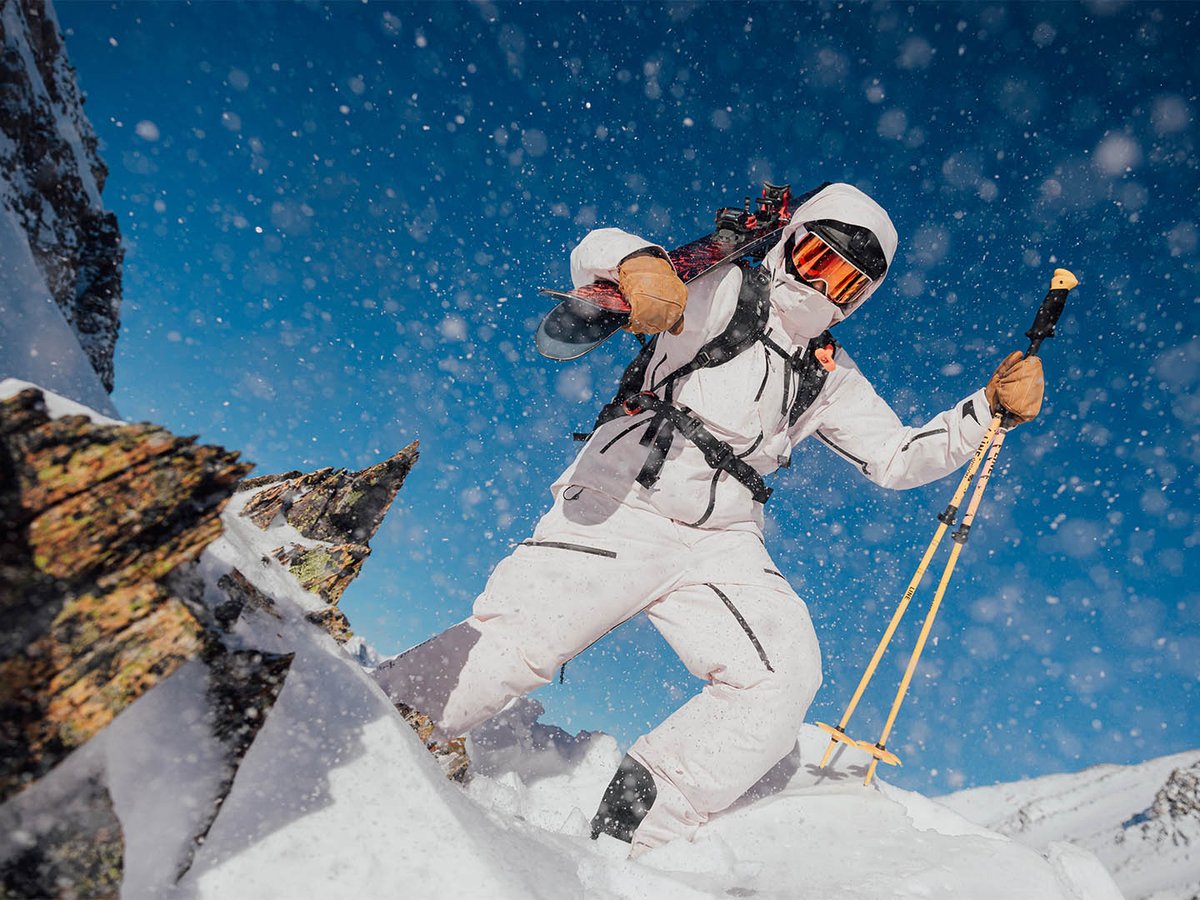 Skier walking holding skis and poled on rocly snow mountain top with view from below.