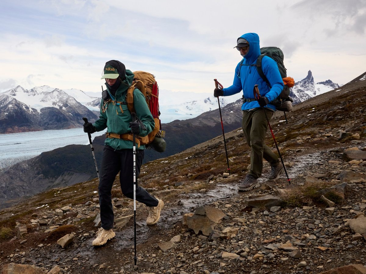 Two backpackers with trekking poles hiking a wet, rocky mountain trail above a glacier and snow-capped peaks.