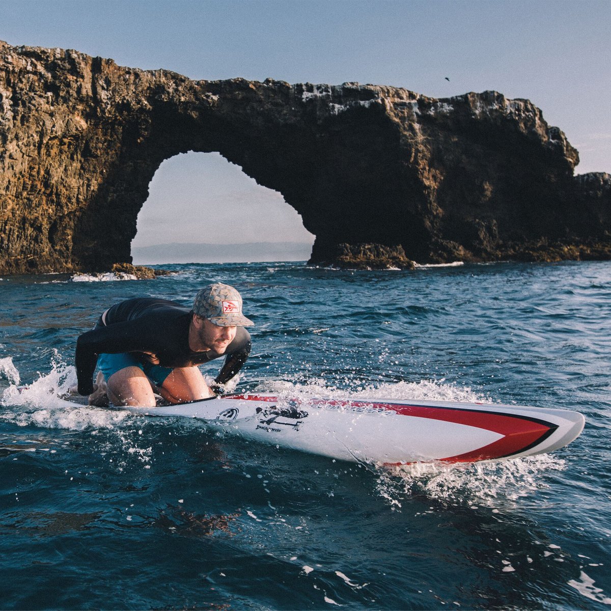 Man surfing on a white and red surfboard in ocean waves with a dramatic natural rock arch formation in the background.