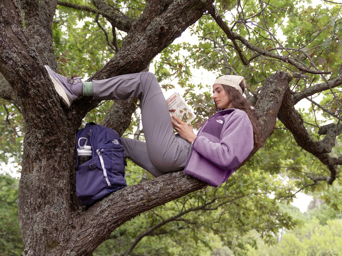 A teenager lounging on a tree branch with a book and a backpack.