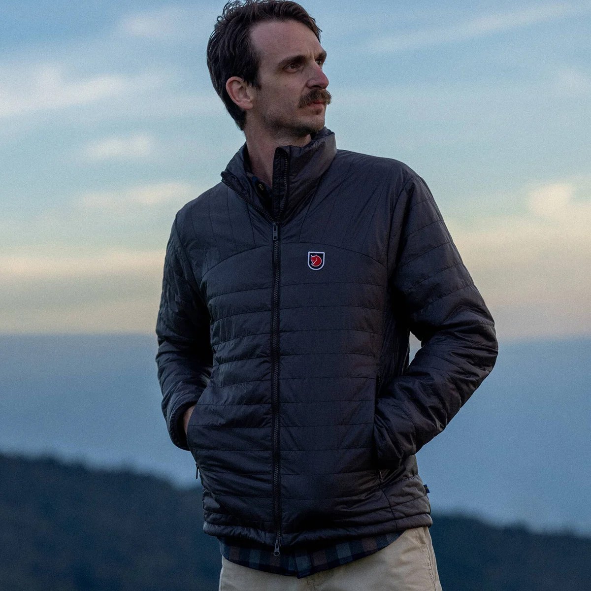 A man wears the Fjallraven Expedition jacket while standing against a gray twilight sky.