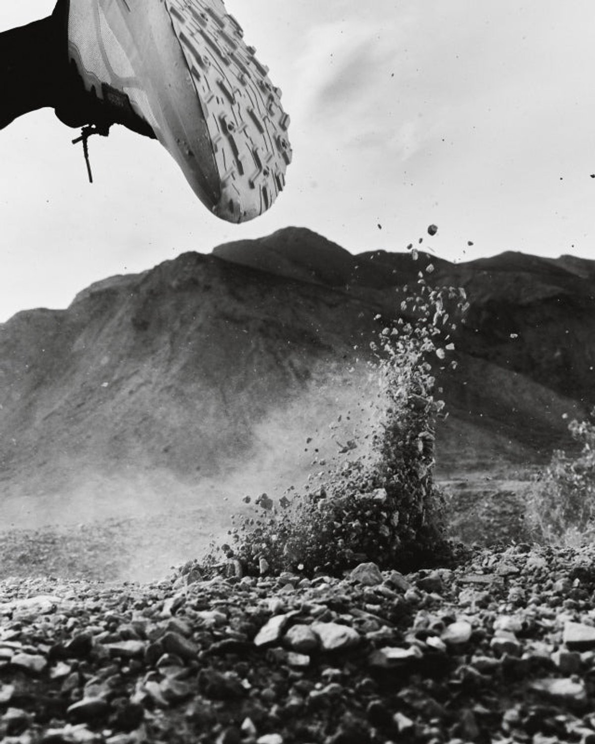 Black-and-white ground-level shot of a Norda outsole overhead as gravel explodes upward, with a desert mountain ridgeline in the background.