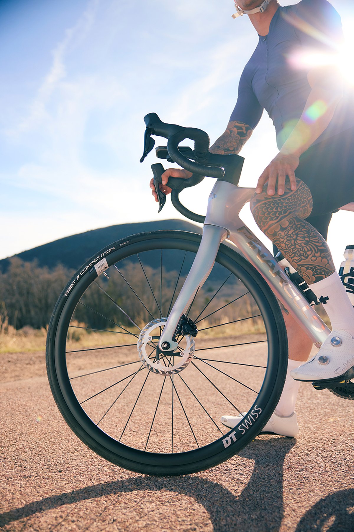 Road cyclist wearing cycling jersey and bib shorts straddling Pinarello road bike with DT Swiss carbon wheelset and disc brake on sunny paved road