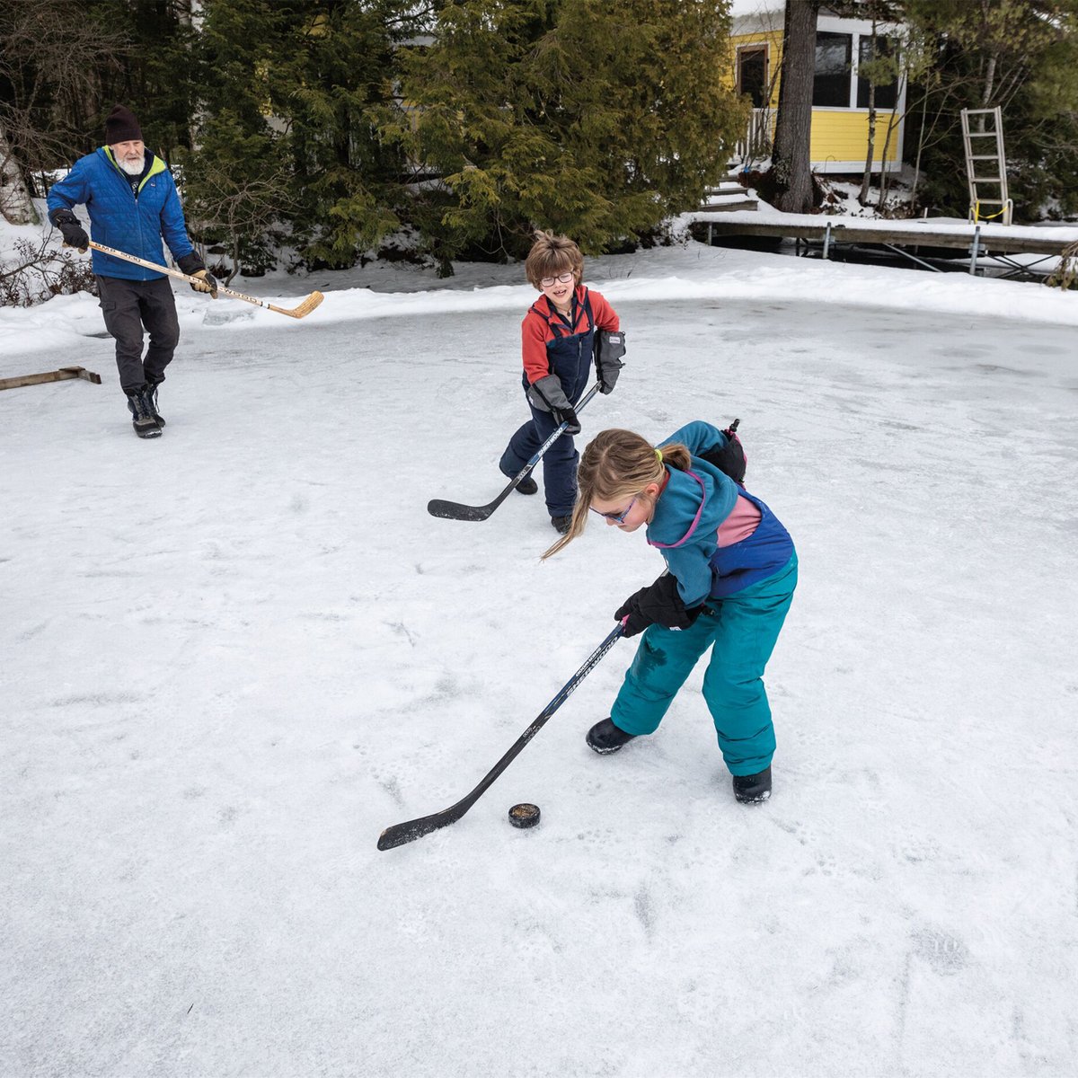 Family playing ice hockey on a frozen lake with an adult and children using hockey sticks and a puck during winter outdoor recreation.