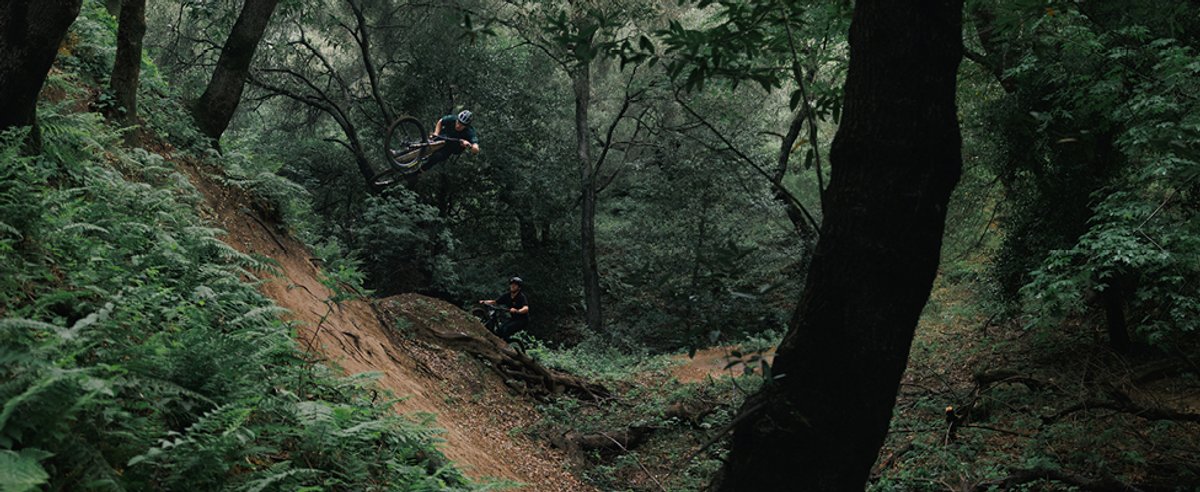 Mountain biker threading tight wooded singletrack trail through dark dense forest environment while another gets big air.