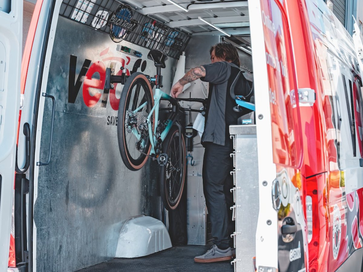 Mechanic working on a teal gravel bike mounted in a repair stand inside the open rear of a mobile service van.
