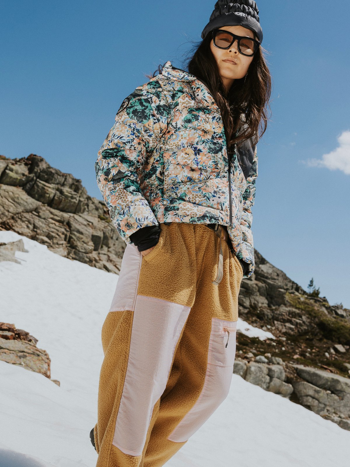 A woman wearing Wander Fleece Joggers and a floral Stansbury ALLIED Down cropped puffy walks across a snow field.