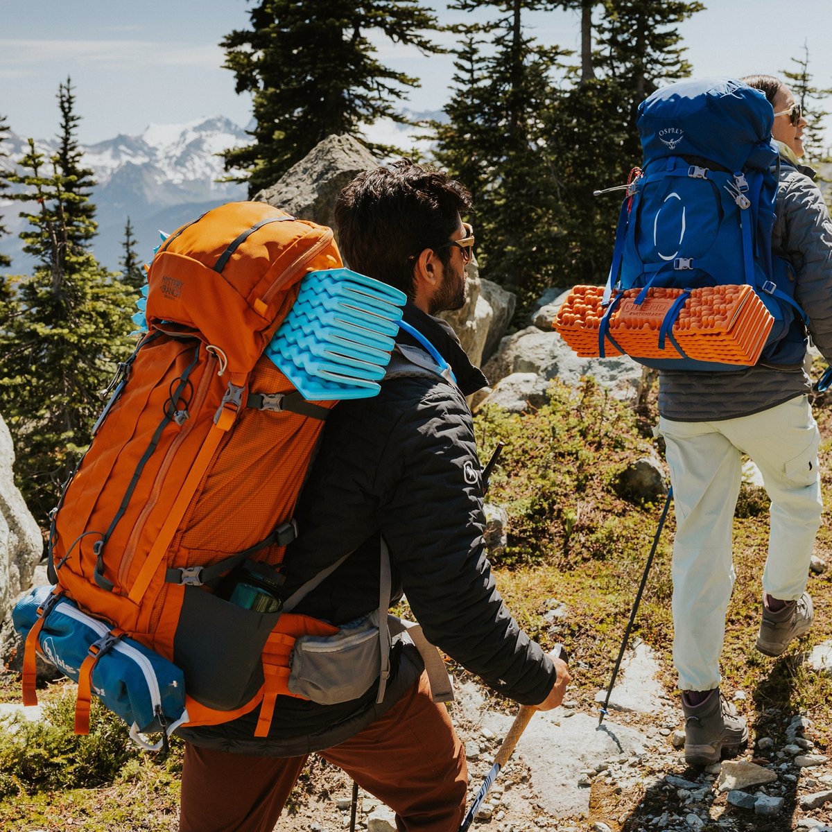 Two backpackers with blue and orange packs hike a high alpine trail.