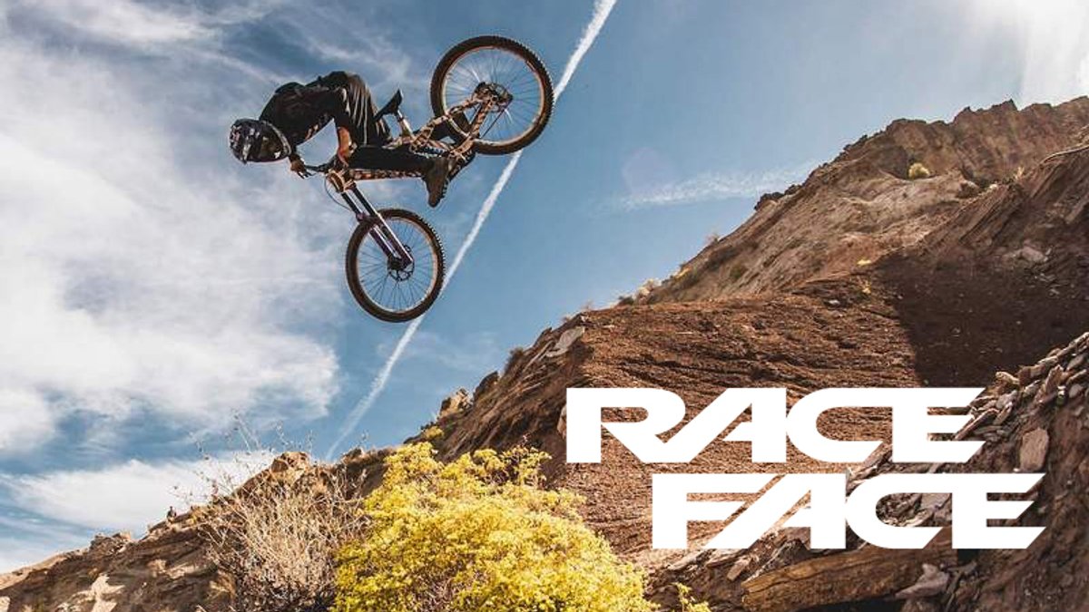 A mountain biker performs an aerial backflip trick off a rocky desert cliff against a bright blue sky.