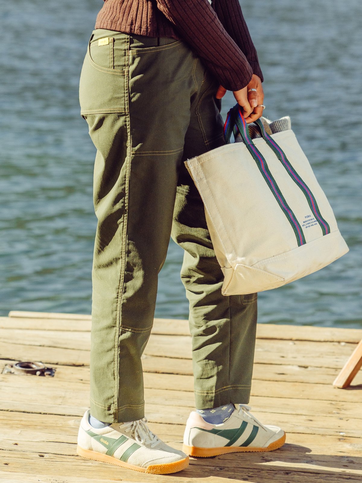 A person stands on a wooden dock by the water, wearing olive green pants, a brown ribbed sweater, white sneakers with green stripes, and holding a large cream-colored tote bag with blue and green striped handles.