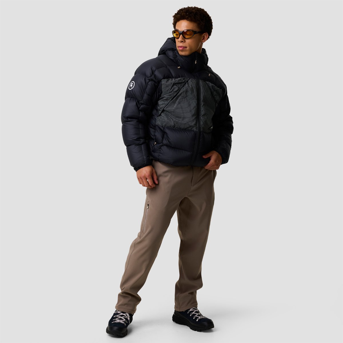 Man wearing a black puffer jacket with built-in backpack straps, tan cargo pants, and black hiking boots, styled against a neutral background.