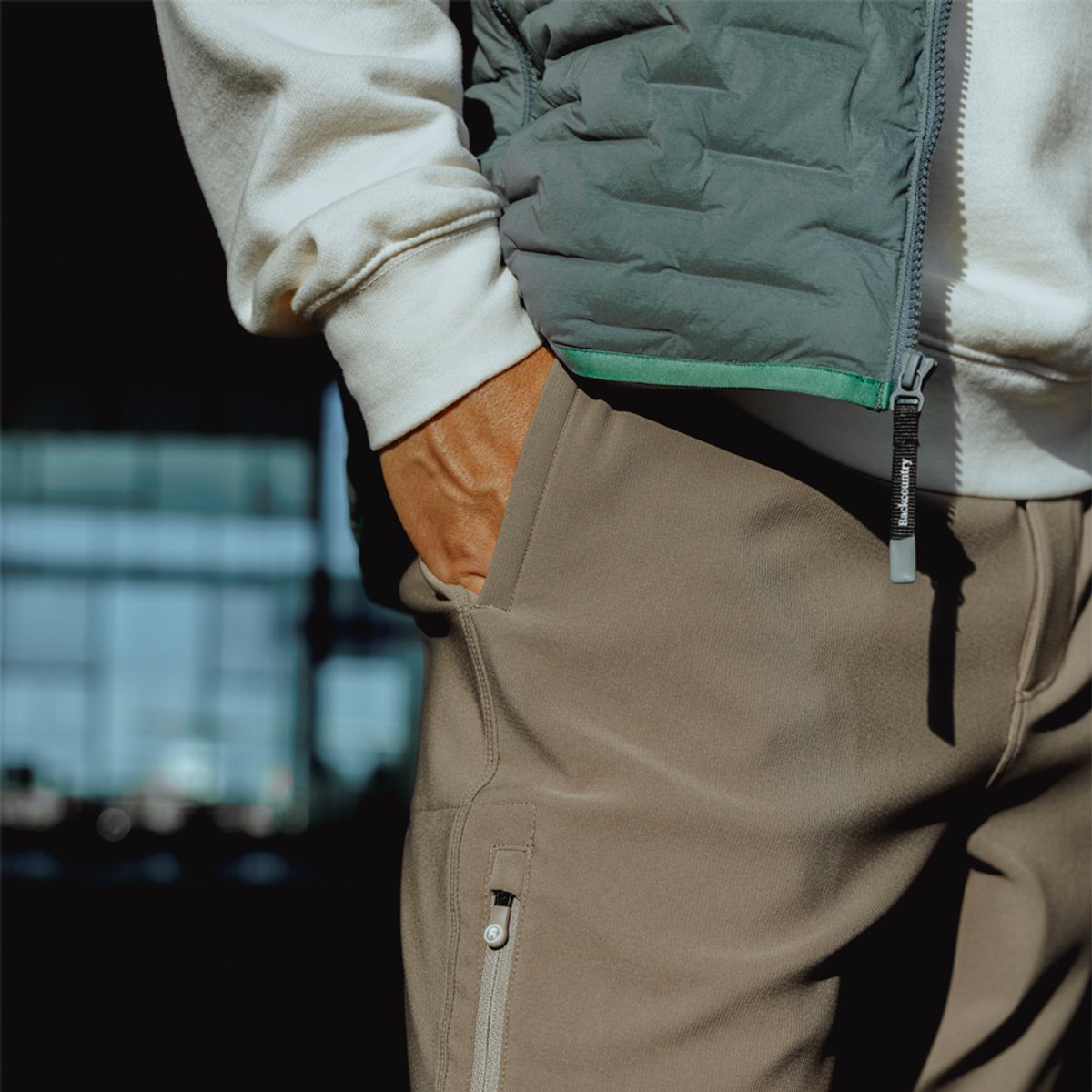 Man wearing a sage green puffer jacket over a cream sweater with beige cargo pants featuring functional pockets and zipper details.