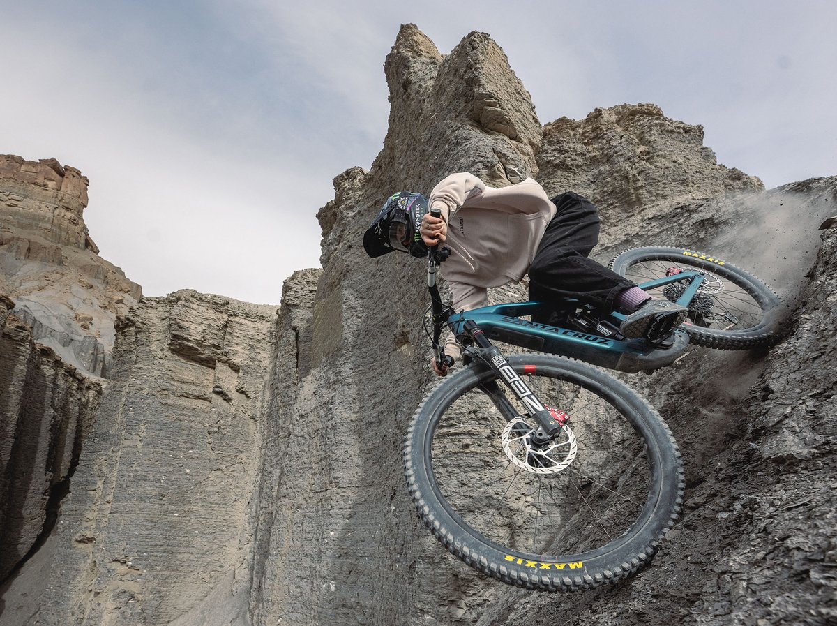 Rider carves a near-vertical dirt wall on a teal Santa Cruz mountain bike, Maxxis tires biting into loose terrain below jagged rock spires.