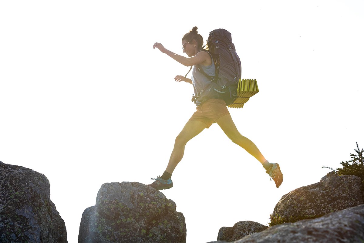 A woman jump from rock to rock while wearing an Osprey backpack.