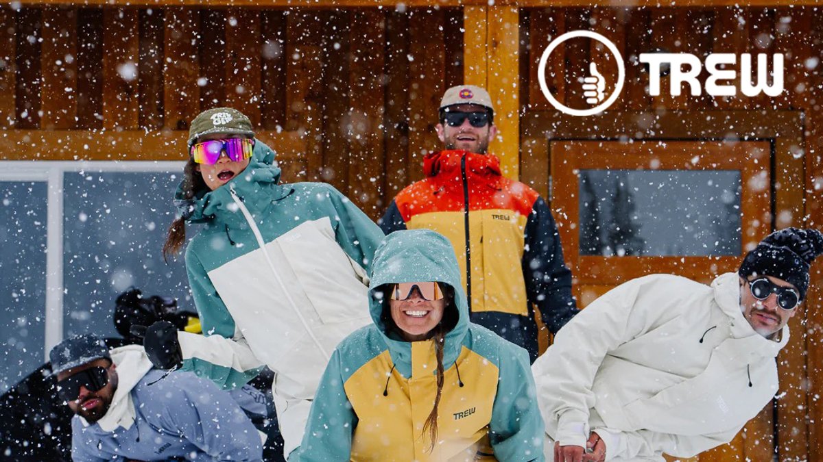 Group of snowboarders and skiers wearing colorful TREW winter jackets and snow gear posing outdoors in heavy snowfall during winter sports conditions.