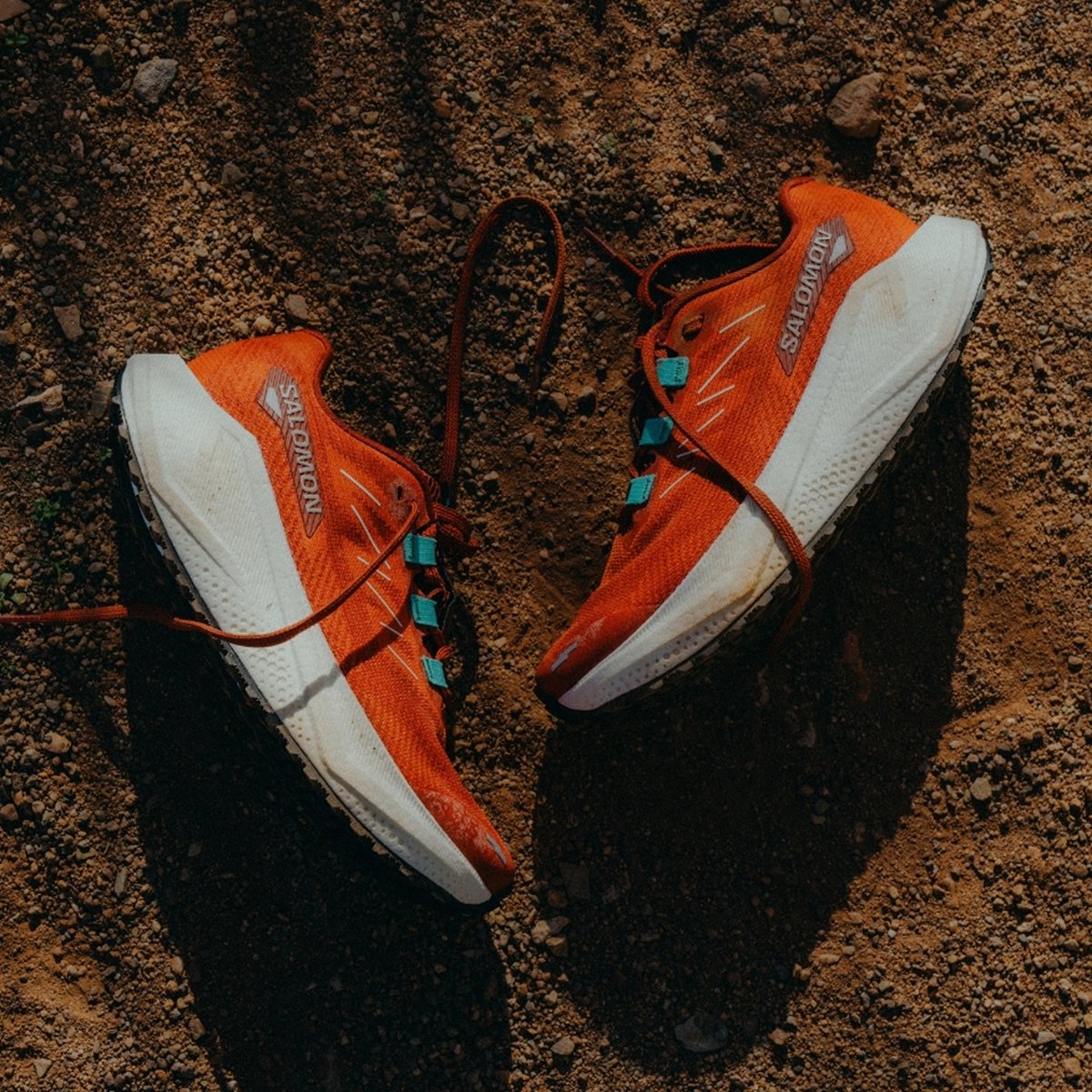 A pair of orange Salomon running shoes with white soles and teal lace loops are lying on a patch of brown dirt, with the untied laces loosely spread around the shoes.
