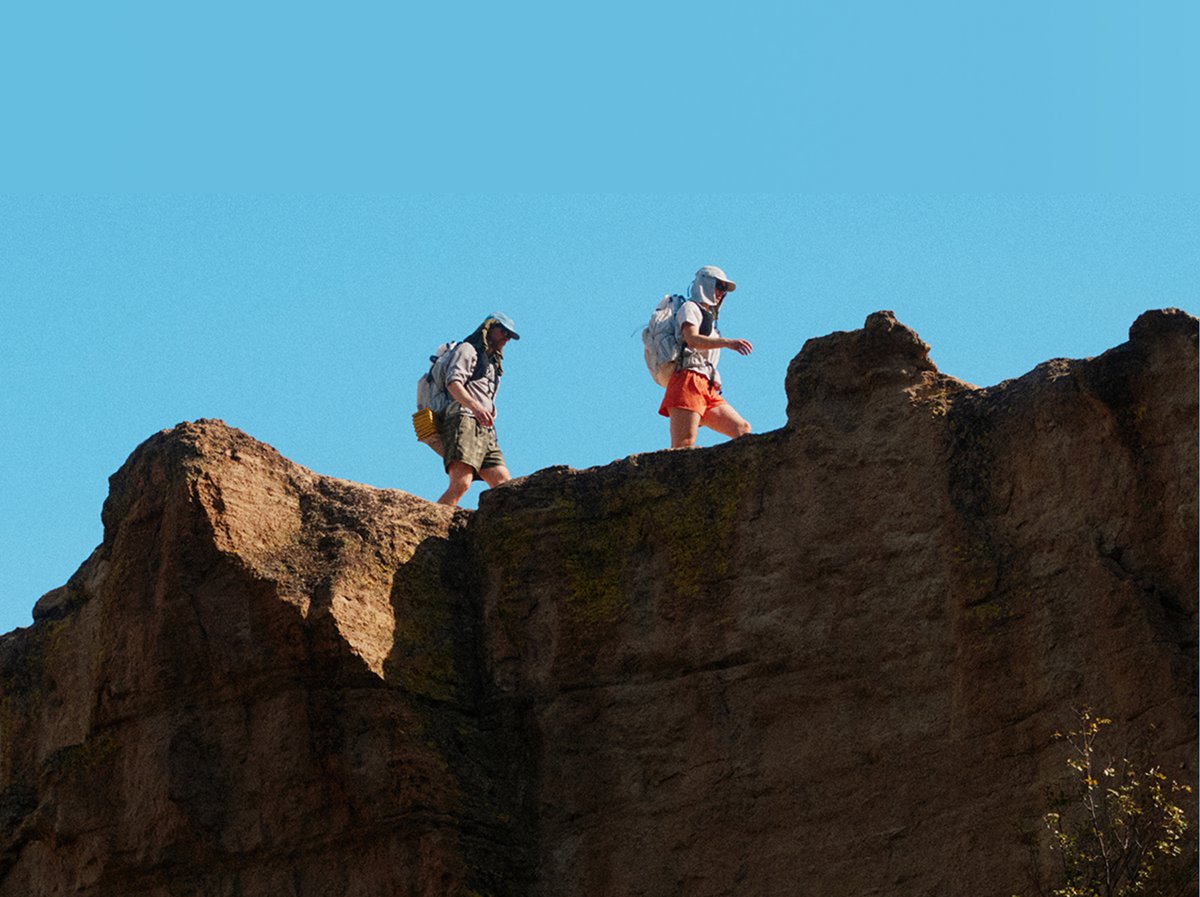 Two hikers traversing a sun-exposed sandstone ridgeline with loaded packs against a clear blue sky.