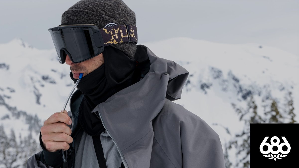 A snowboarder wearing a grey technical jacket and goggles drinks from a hydration tube with snowy mountains in the background.