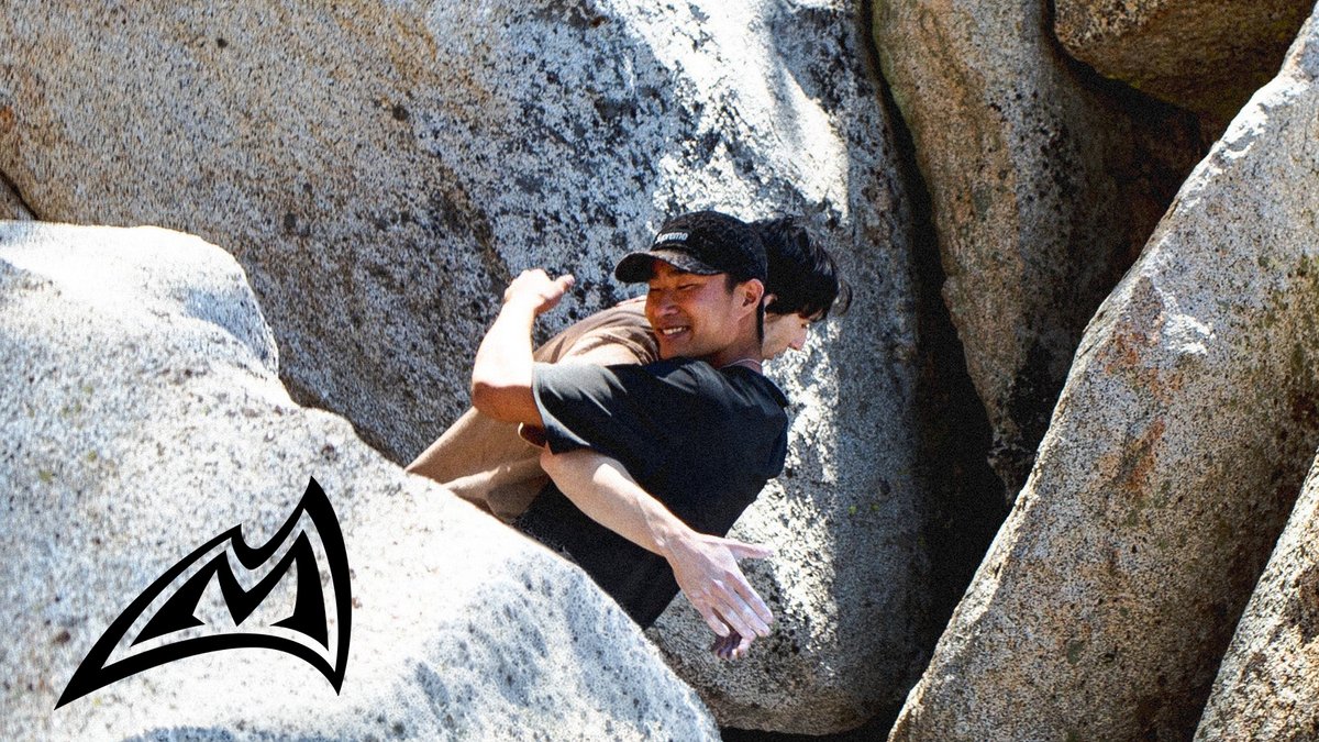 Man rock climbing between large granite boulders wearing black athletic shirt and cap during outdoor bouldering adventure.
