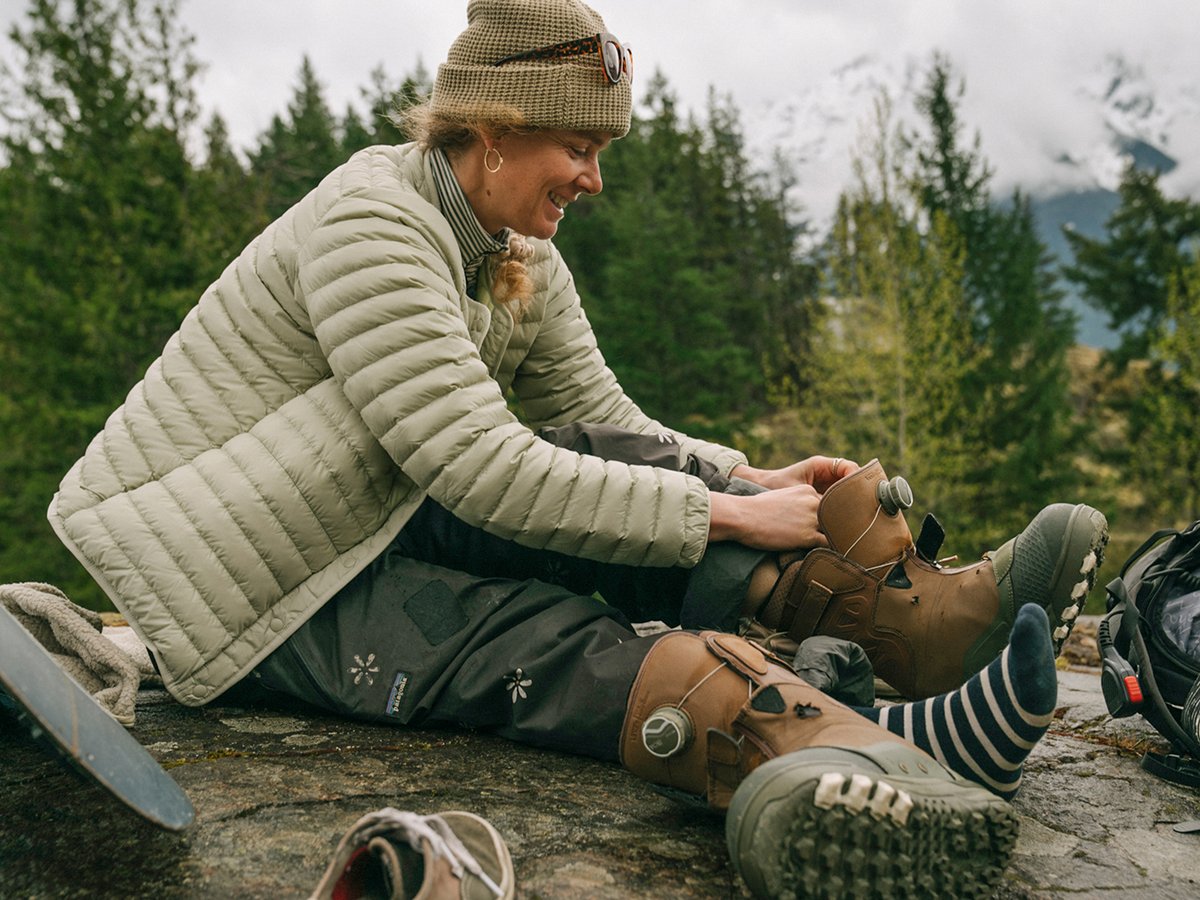 Woman lacing up snowboard boots on a rocky ledge wearing an insulated jacket, surrounded by evergreen trees and mountain views